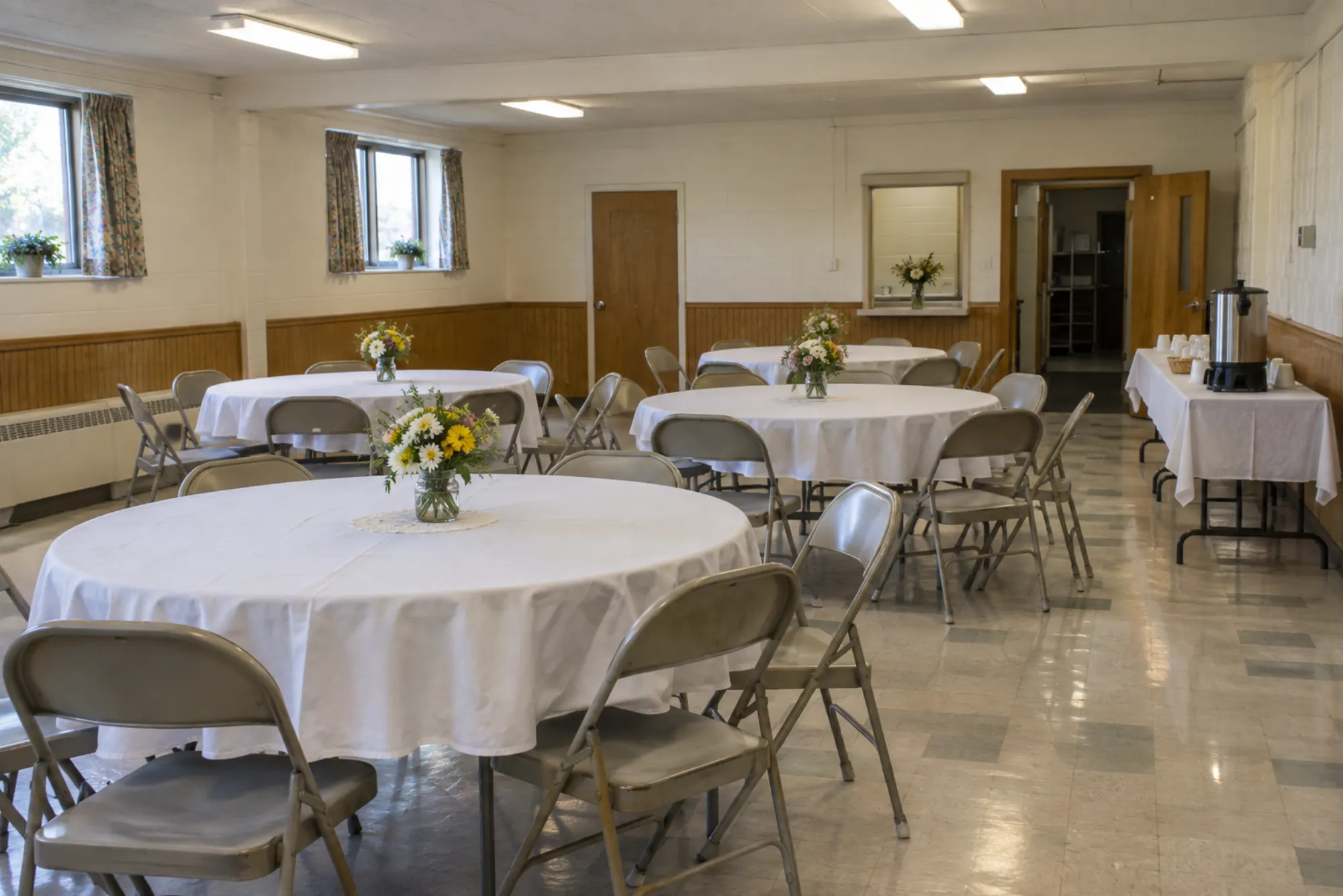Round tables in white cloths with simple flowers in the community hall, a coffee urn on the side table